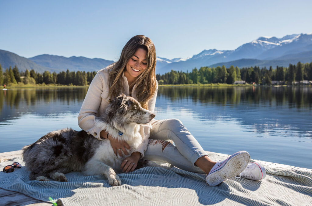 A woman sits with her dog on the shores of a lake in Whistler.