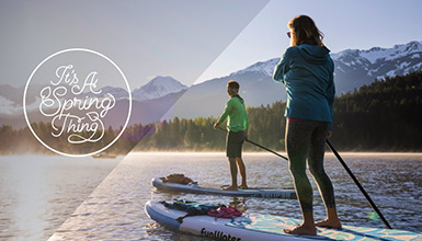 Two paddleboarders glide across a misty mountain lake at sunrise, framed by snow-capped peaks. A golden glow illuminates the water, capturing the thrill of spring adventure in Whistler. Overlay text reads 'It's A Spring Thing' in elegant script.